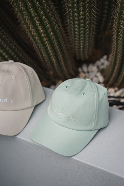 Two baseball caps, one beige and one light green, on a surface with a cactus in the background.