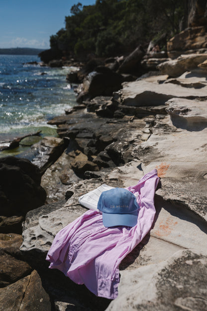 Pink shirt and blue ilk studio cap on a rocky beach with ocean and trees in the background