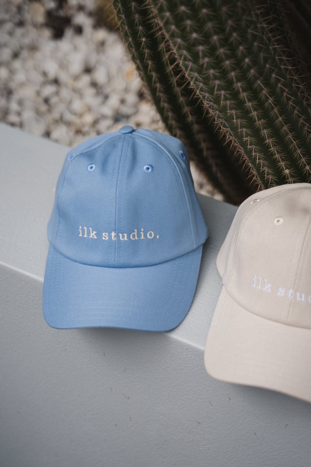 Two baseball caps, one blue and one beige, with 'ilk studio.' text, against a natural background.